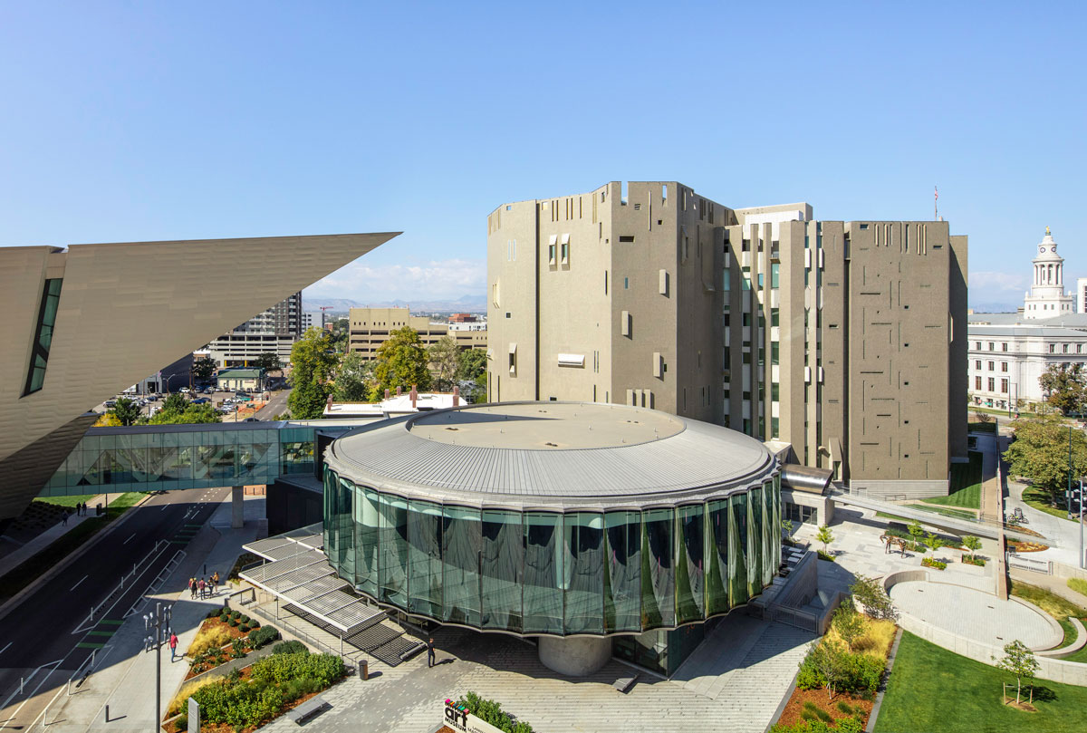 A view of the Denver Art Museum's three buildings on a light-blue skied summer's day. The triangular point of the main building sits above a round glass building with another geometric five-story building behind it.