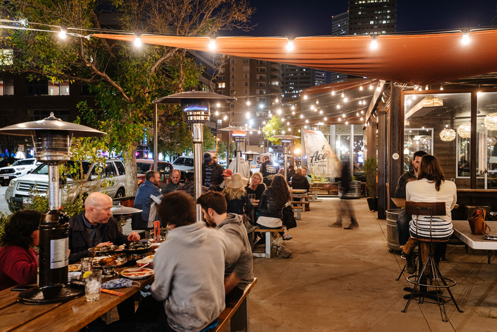 Diners sit at sleek, dark-wood tables in a closed-off courtyard space outside a white-brick building in Denver, Colorado. Tables are filled with glasses of water and orange juice and plates of food.