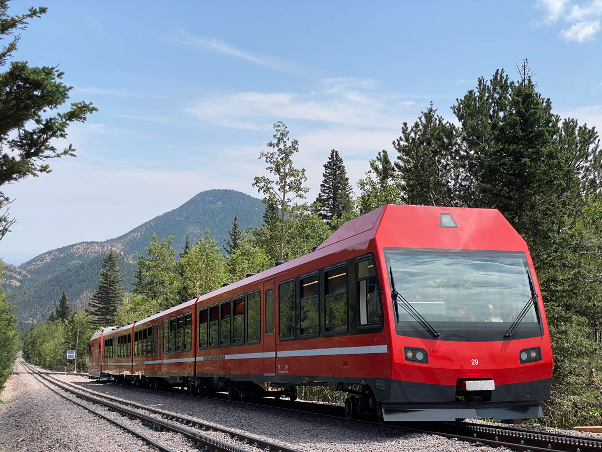 A boxy, modern, red train with windshield wipes on the front cruises on a track through a forest near Pikes Peak, Colorado, with mountains in the background
