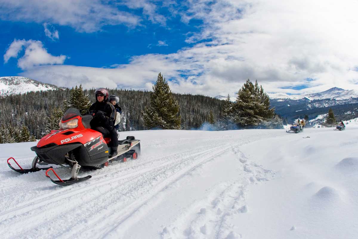 Two snowmobiles, each with two riders cruise through a snowy fields surrounded by thick, fluffy pine trees and tall mountain peak in Colorado's backcountry.