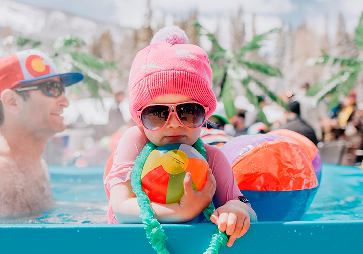 The coolest kid ever hanging out in a pool with sunglasses and a pink cap.