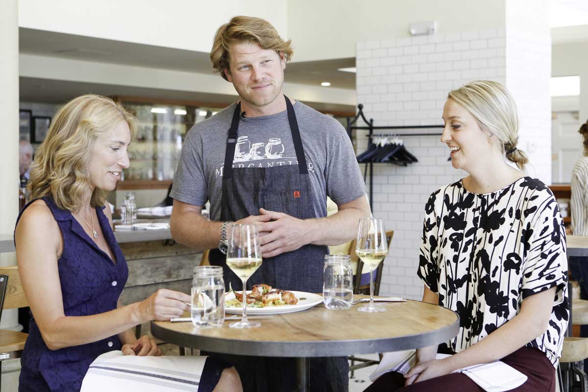 Chef Alex Siedel stands next to two blond women with full plates and glasses full of white wine