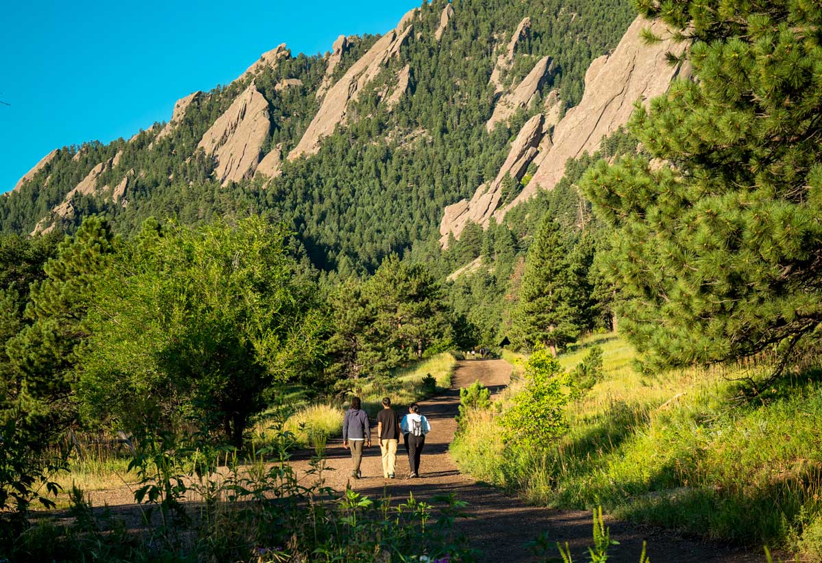 Three people dwarfed by sloped granite peaks walk along a dirt trail