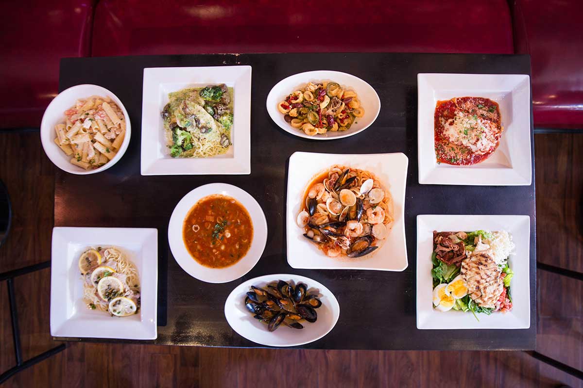 An aerial view of a table filled with white square and round dishes holding a variety of pasta and seafood