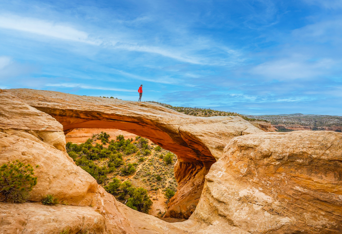 Someone stands on the Cedar Tree Arch in Rattlesnake Canyons in McInnis National Conservation Area in Grand Junction, Colorado. The arch is massive and looms over desert-looking landscape. The sky above is blue full of wispy clouds.