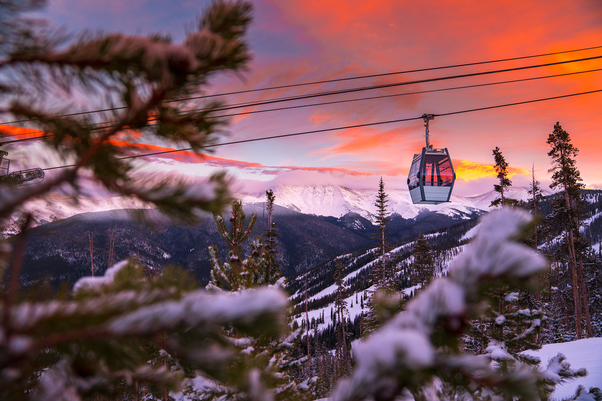 A picturesque image of the Winter Park gondola inching down above snow-covered and pine-peppered hills. The sky above is panted in deep, vibrant oranges and pinks of sunset, and the mountains in the background are covered in snow. In the foreground and to the left of the photo are out-of-focus pine tree branches.