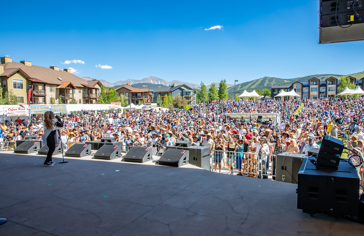 A crowd of people face a stage on which a performer sings. There are mountains behind the crowd.