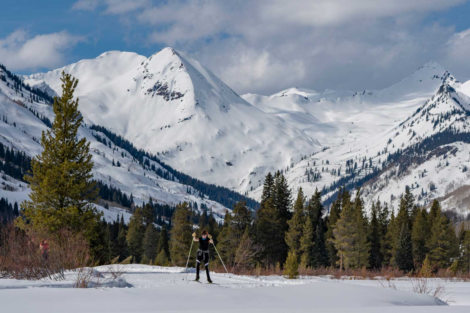 A cross-country skier glides through a snow-covered meadow with snowy peaks in the background