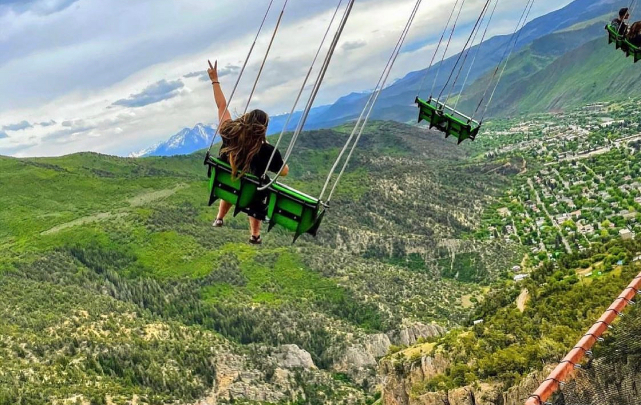 A picture shot from behind of a person in a swing making a peace sign as they soar over a lush canyon on the Glenwood Caverns Flyer at Glenwood Caverns Adventure Park in Glenwood Springs, Colorado.