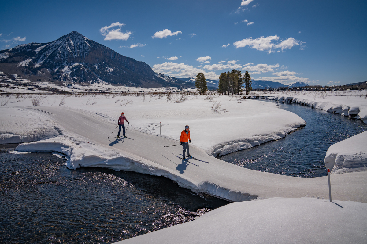 Two people cross-country ski on a snowy trail crossing a running creek in Crested Butte and Gunnison, Colorado