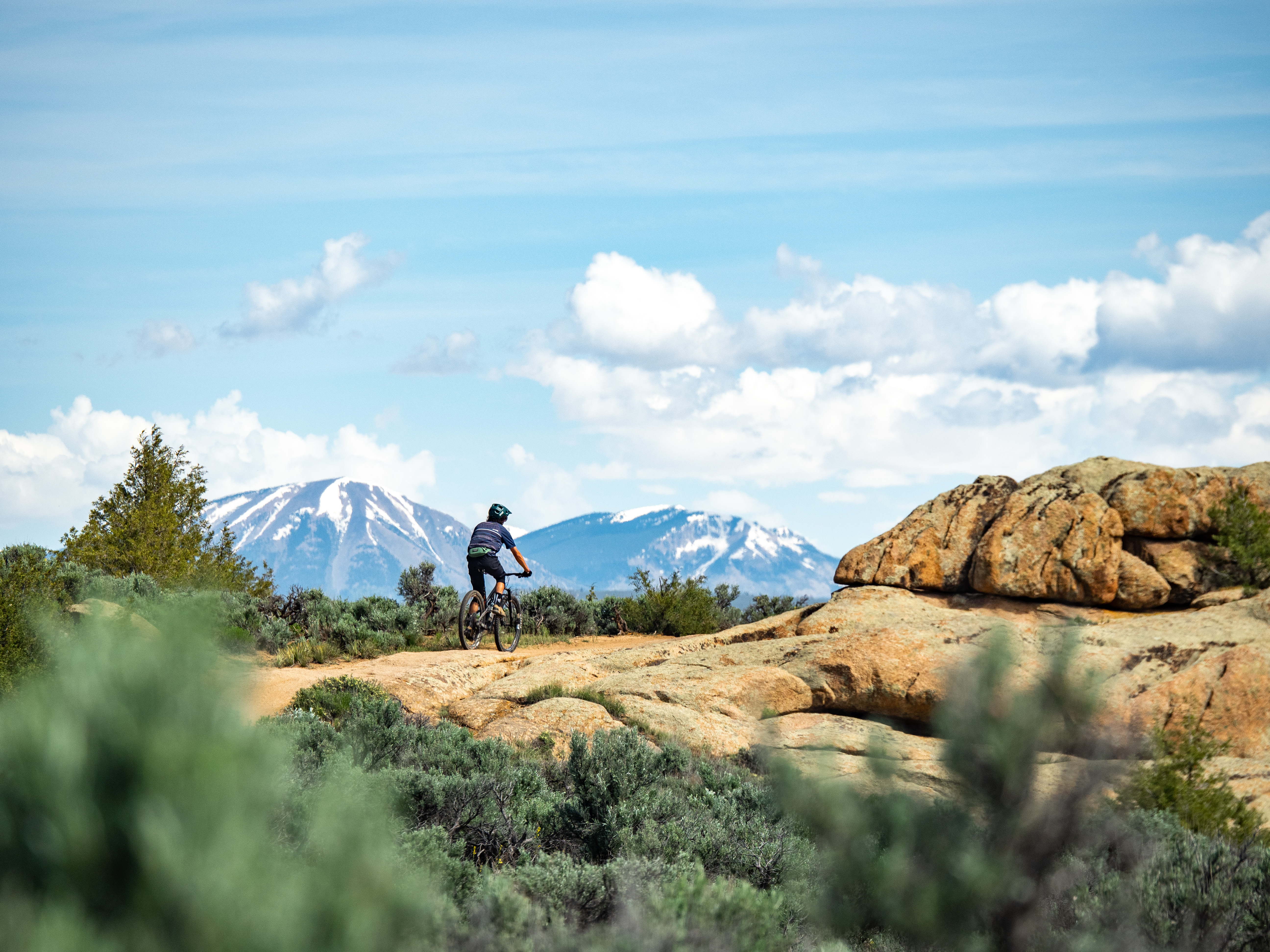 A mountain biker rides a trail in Gunnison, Colorado with snowcapped mountains in the background.