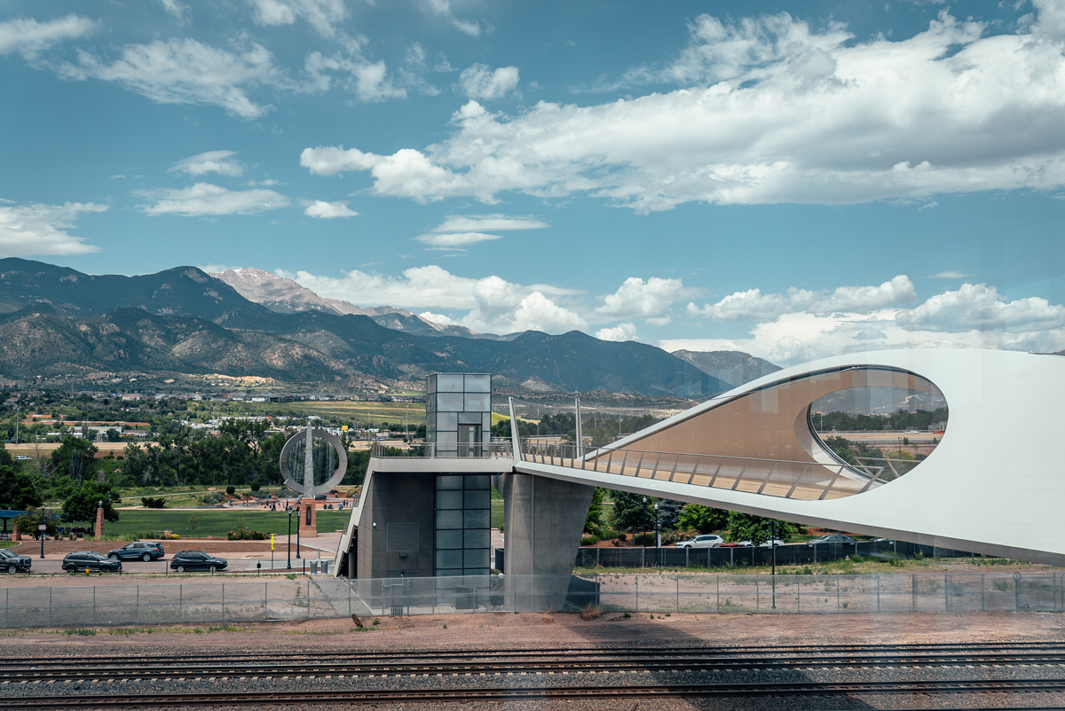 A futuristic bridge over train tracks leading to a park with a large silver sculpture with water feature and Pikes Peak in the background at US Olympic & Paralympic Museum in Colorado Springs