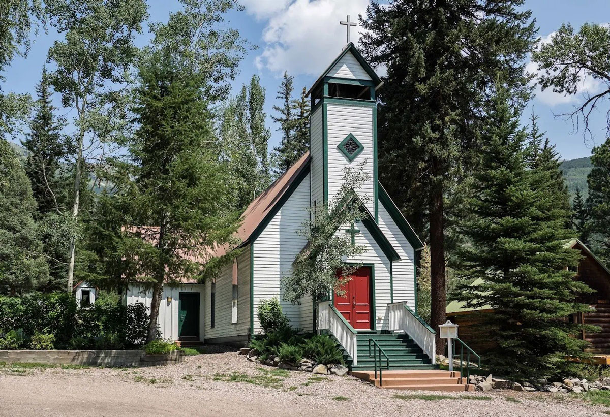 A white church with red doors and green ascent colors, set against mountain peaks and pine trees beside it in Marble, Colorado.