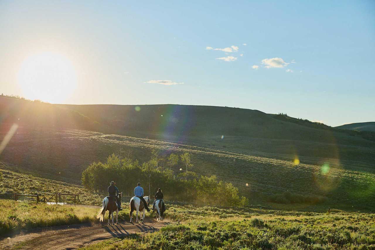 Three riders on horseback ride into a hilly area as the sun sets