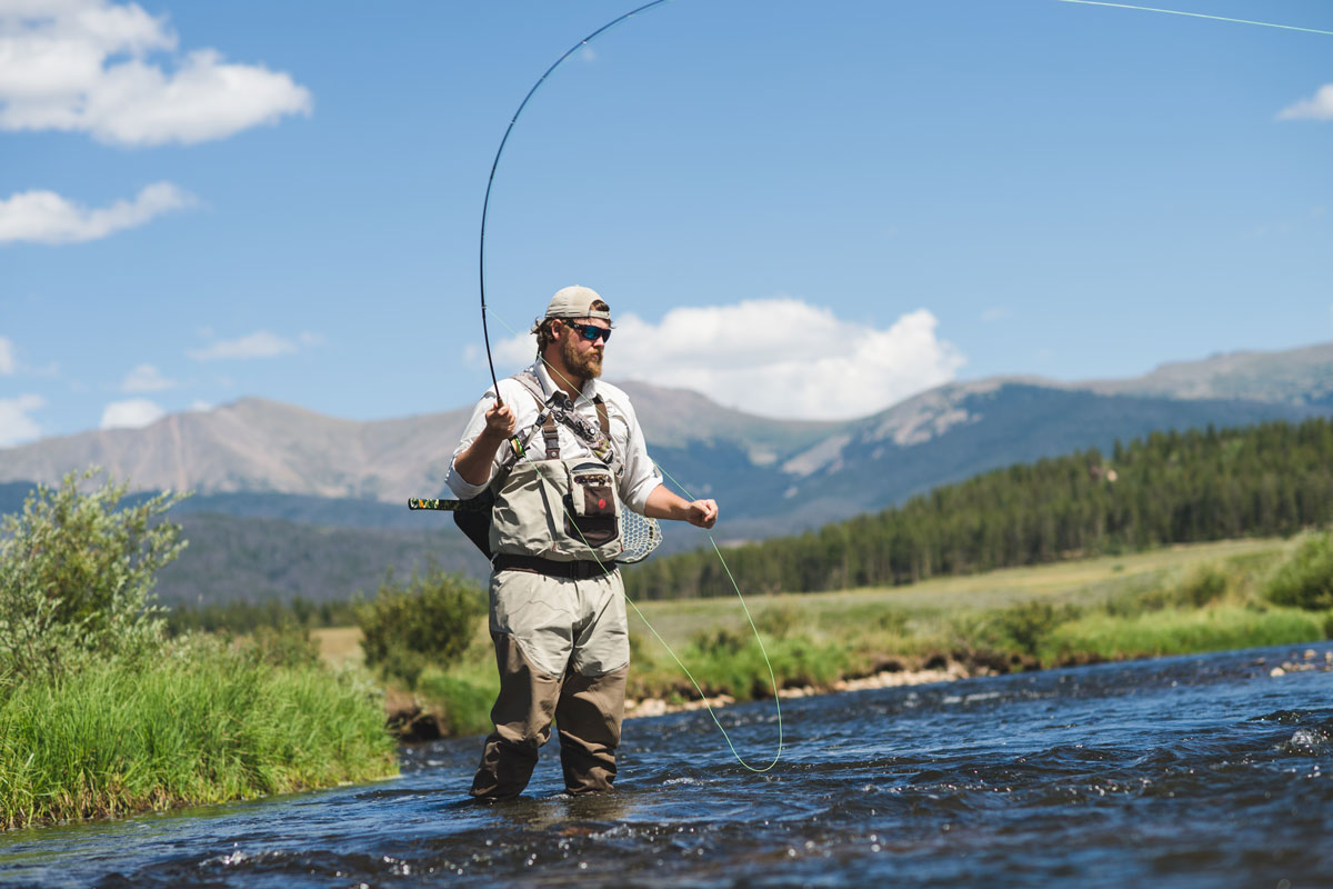 A man fly-fishing in the middle of a river with snowy mountains in the background