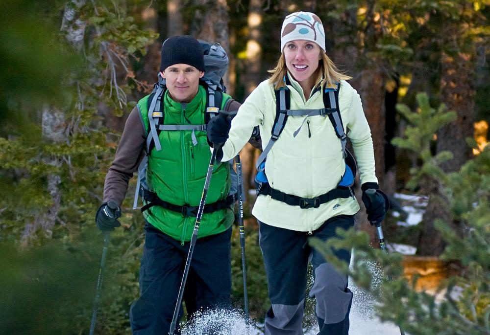 Outside Boulder, Colorado, two winter hikers trek through snow in a pine forest. They wear backpacks and use walking poles as they walk.
