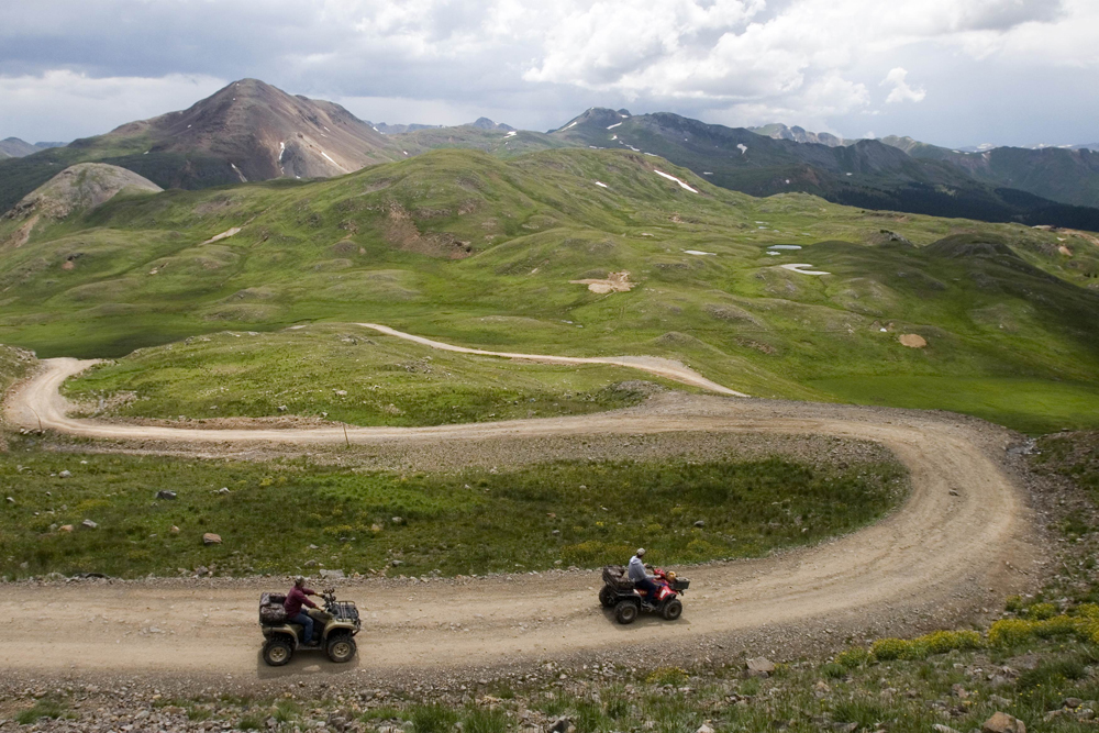 Two red ATVs climb a switchbacking trail along a green mountainside toward a peak's summit