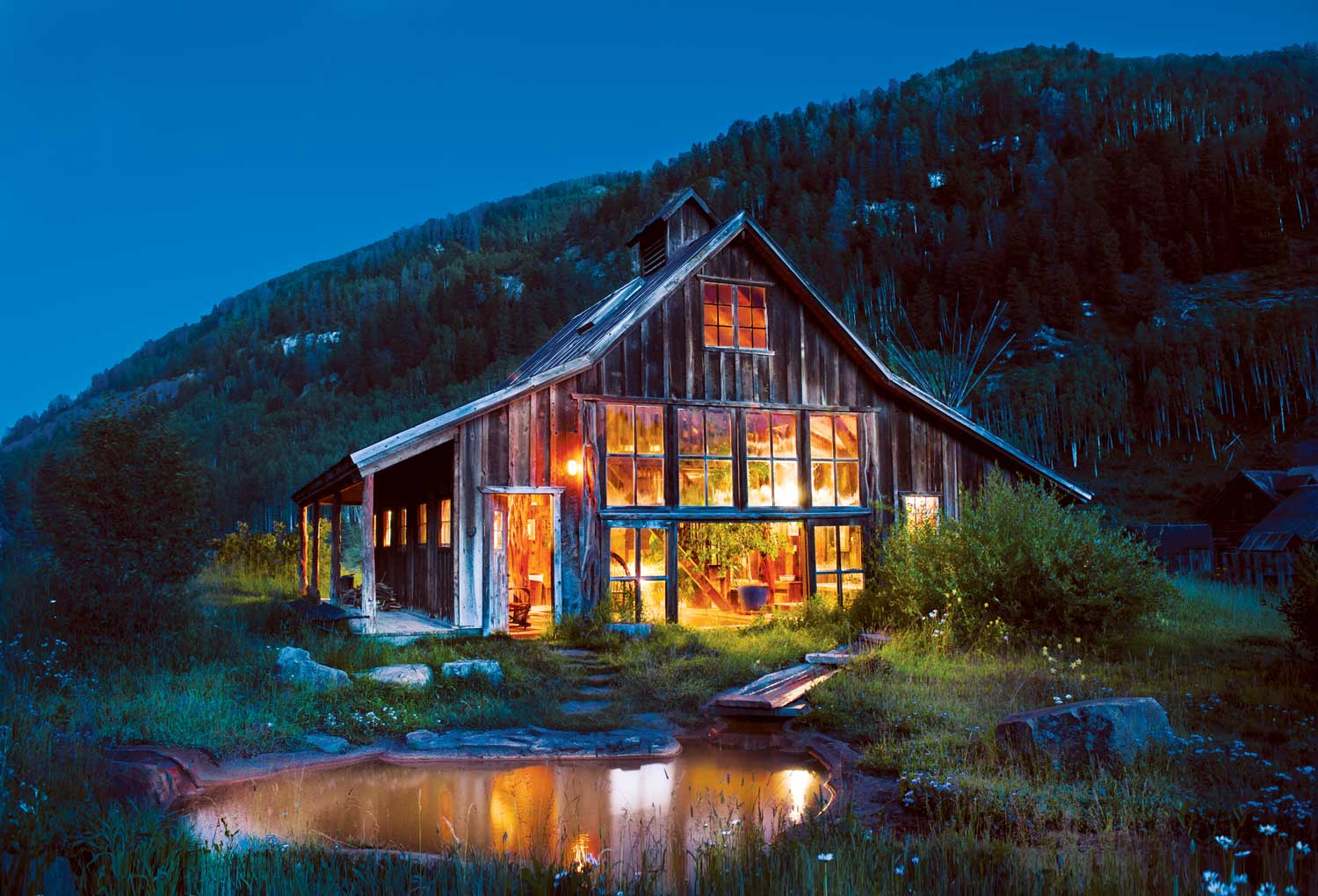 A worn farm building in Colorado glows with golden light against the dark of the evening. A few stone steps away is a small pool of hot water.