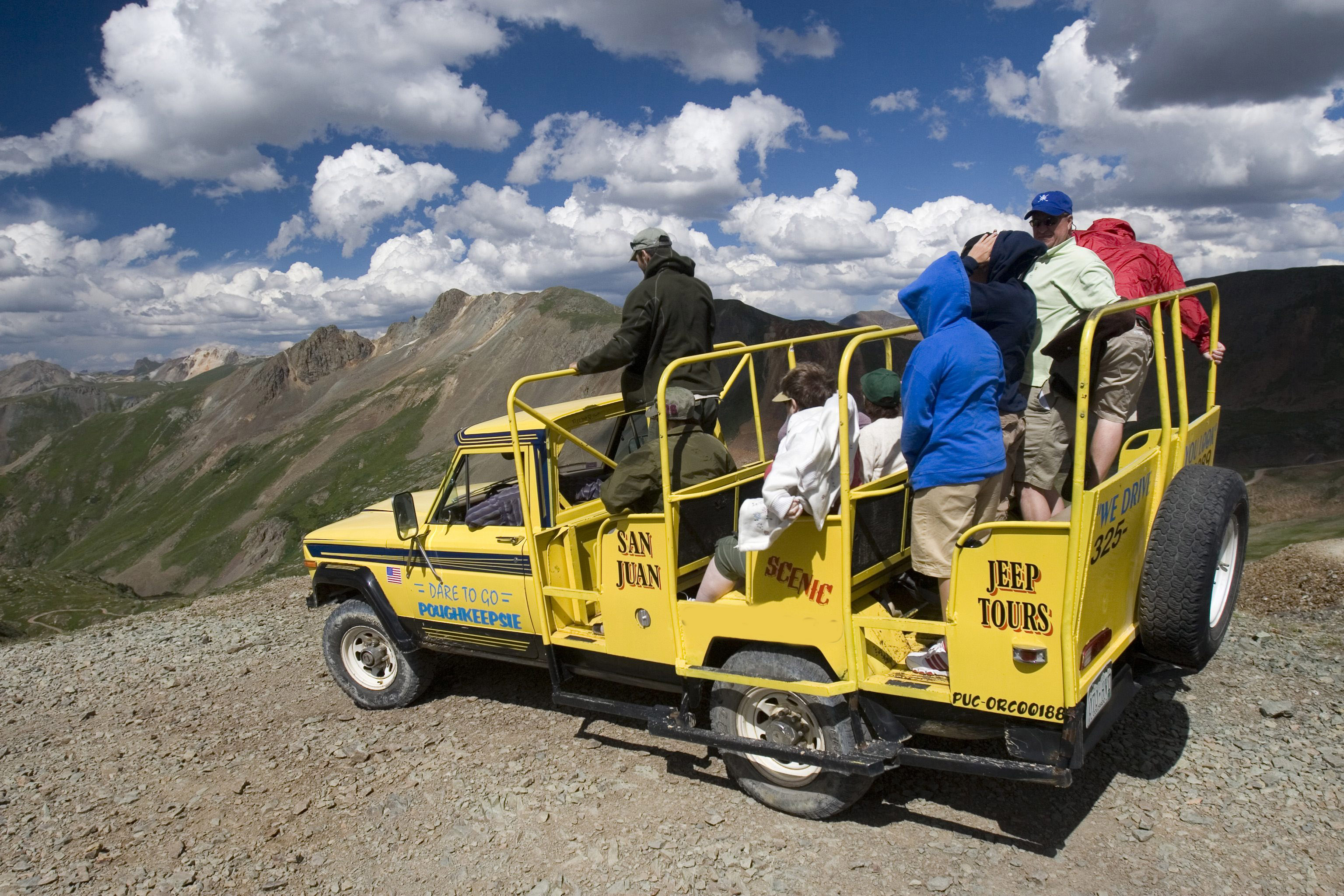 A yellow Jeep modified to fit two rows of tour guests perches on a ledge high in the mountains. The passengers all peer over into the cravasse below