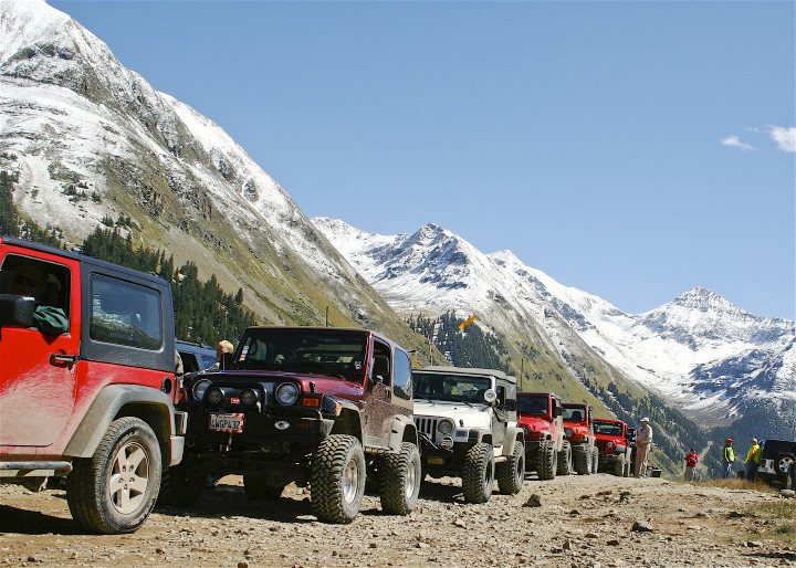 A row of 6 red and white Jeeps with sturdy tires gets ready to brave a rocky road; snowcapped peaks rise in all directions