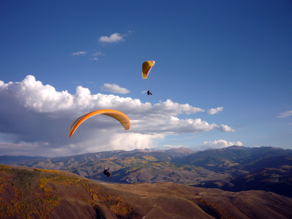 In a mostly blue sky, with a few friendly clouds, we see two paragliders gently moving lower under their orange parachutes into the soft brown foothills of Vail