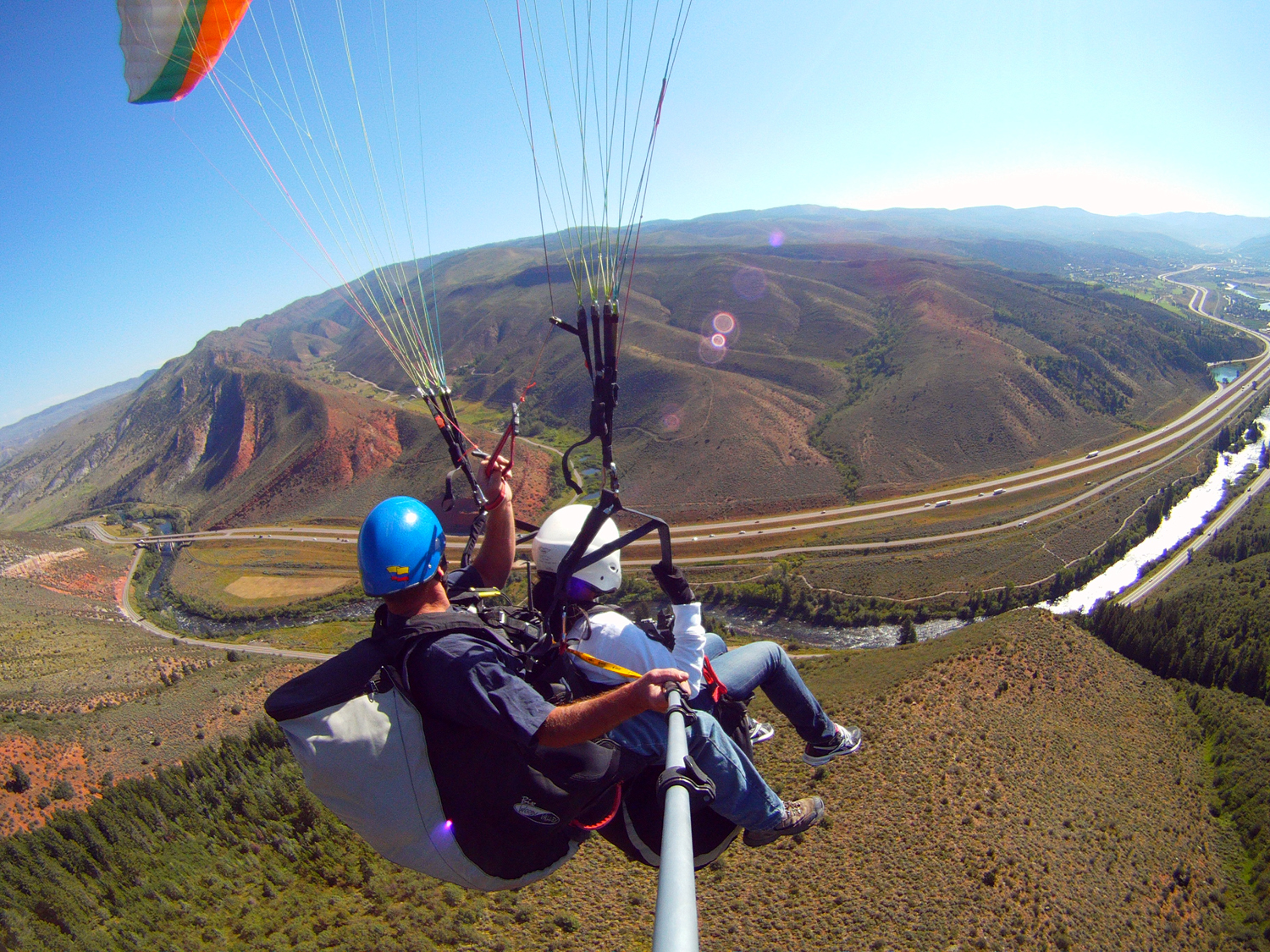 A paraglider working his way gently down to the ground takes a camera using a selfie stick. Below, we see green evergreens and some rolling red-rock hills