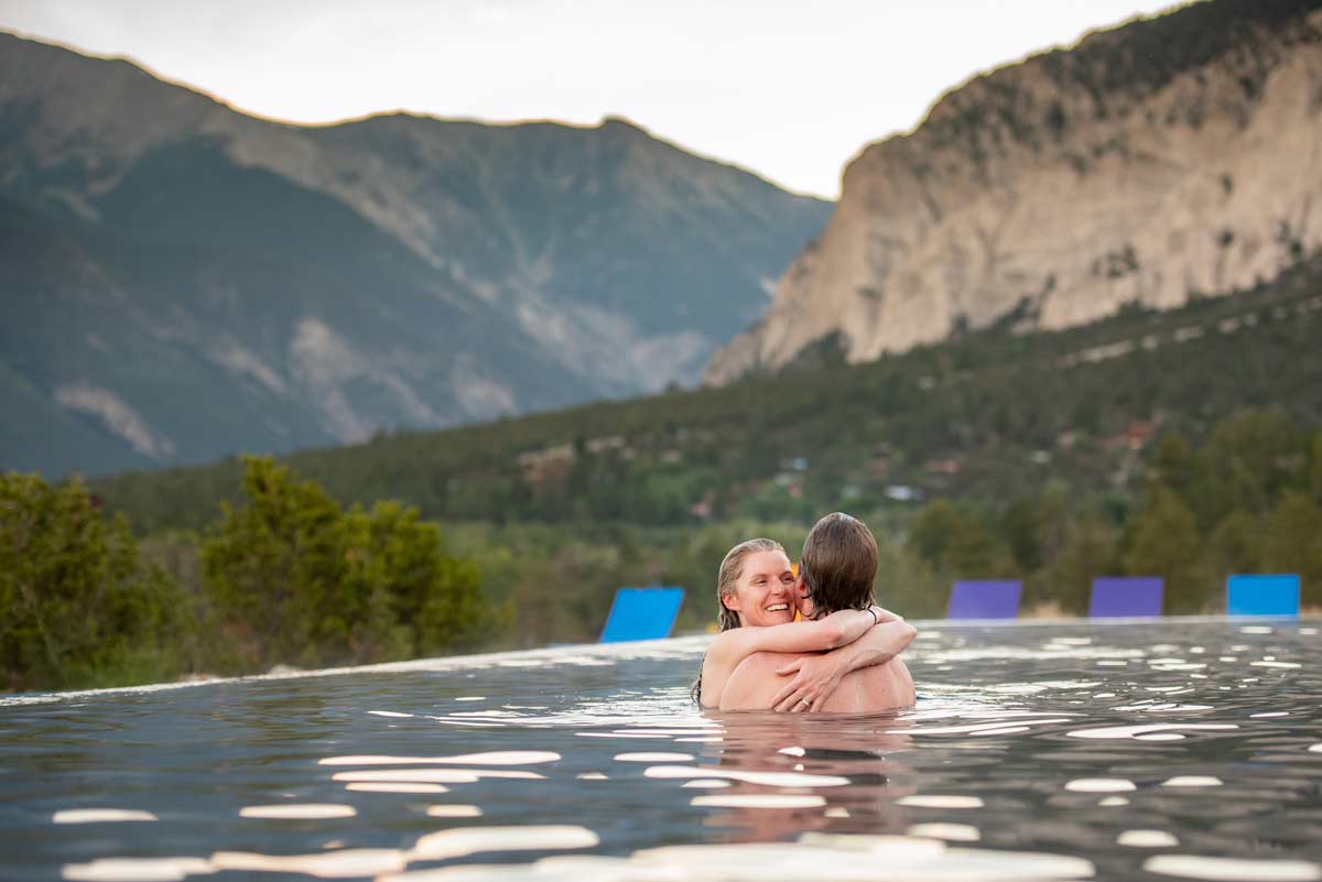 A couple embraces in an infinity pool hot springs with mountains and evergreen trees in the background at Mount Princeton Hot Springs Resort.