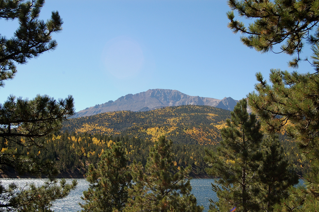 Pikes Peak sits beneath a blue sky with a sparkling lake in the foreground. There are evergreen trees surrounding both sides of the frame and golden leaves are just starting to appear on trees.