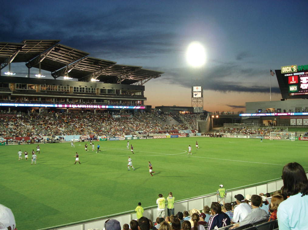 A dusk major-league soccer game with players running around on the field. There's a grandstand with a tilted roof and lots of spectators.