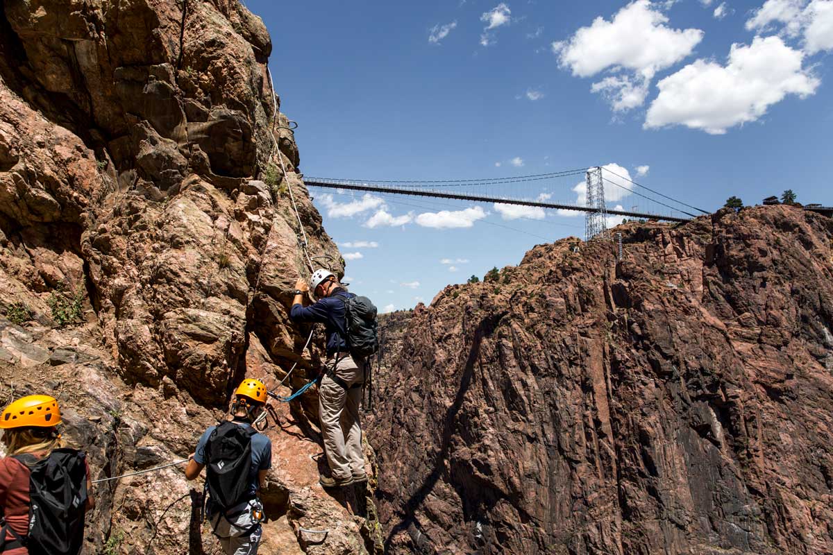 Three rock climbers maneuver and scale a face of the Royal Gorge in Colorado. Overhead, the Royal Gorge Bridge stretches tall and strong across the immense rocky chasm.