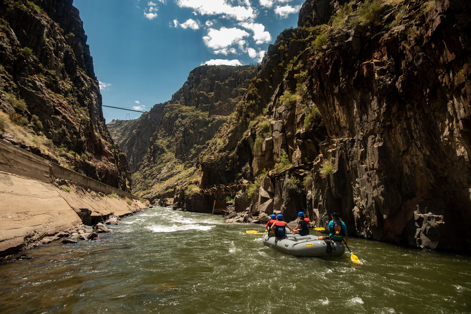 A raft full of paddlers navigates the river while the Royal Gorge Bridge can be seen above the canyon walls