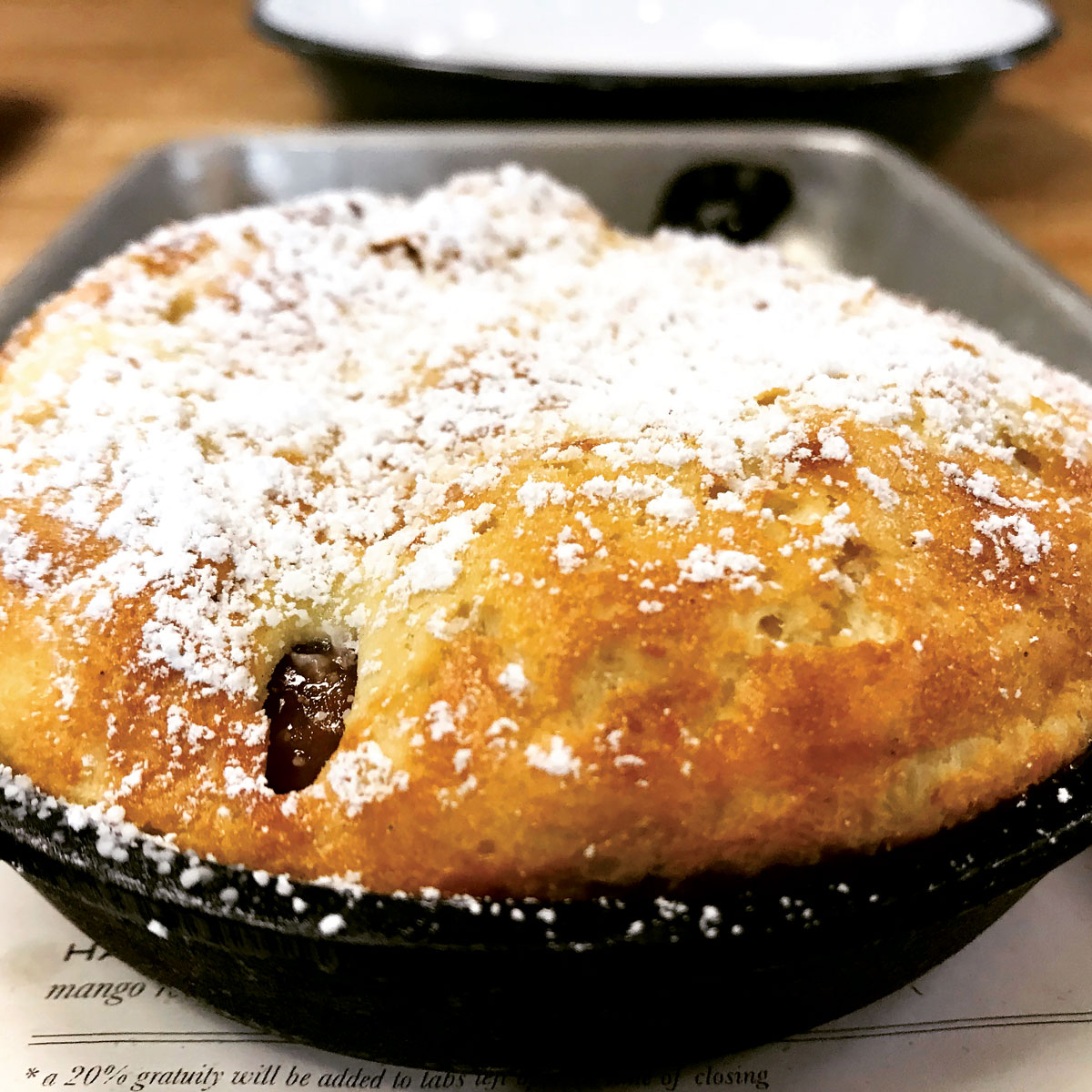 A closeup view shows a fresh pastry cooked to a golden-brown perfection and topped with sweet powdered sugar from Acreage in Colorado. A strategic hole reveals a cherry jam filling.