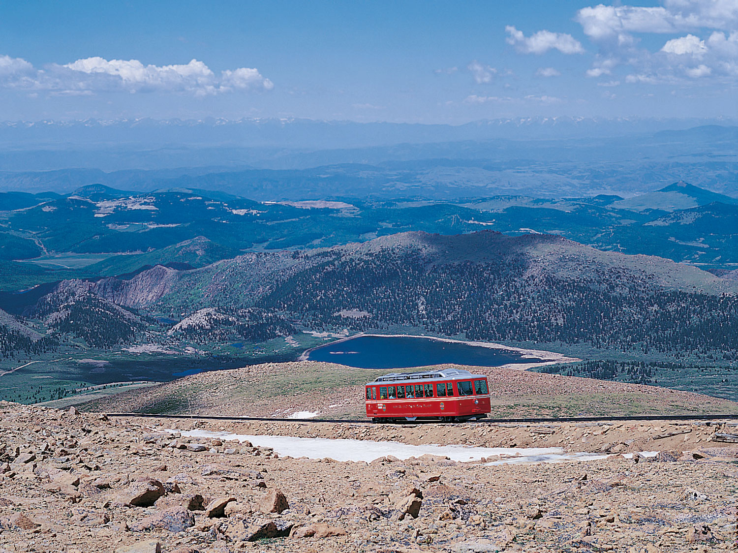 The red car of Pikes Peak Cog Railway makes its way up a rocky incline with snow near the track. In the distance mountains are covered with evergreen trees and hazy clouds.