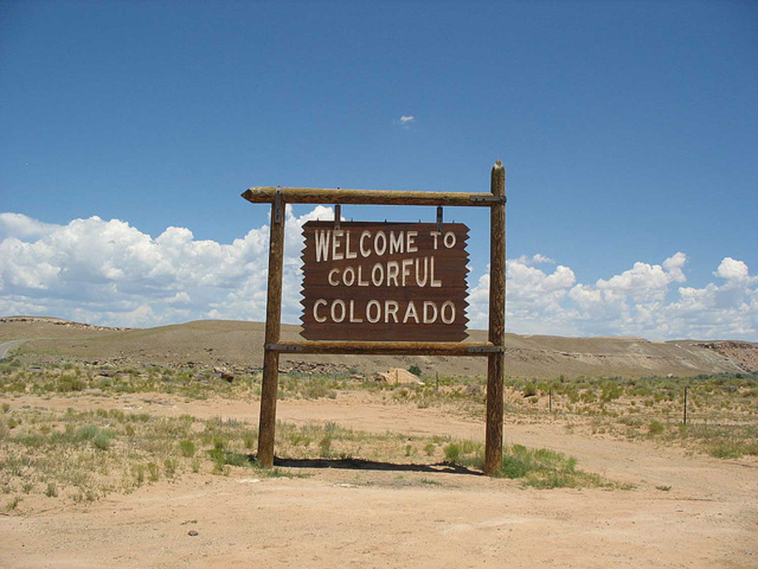A large wooden sign is engraved with the words, "Welcome to Colorful Colorado." Behind the sign are rugged Colorado mountains and a bluebird sky with clouds.