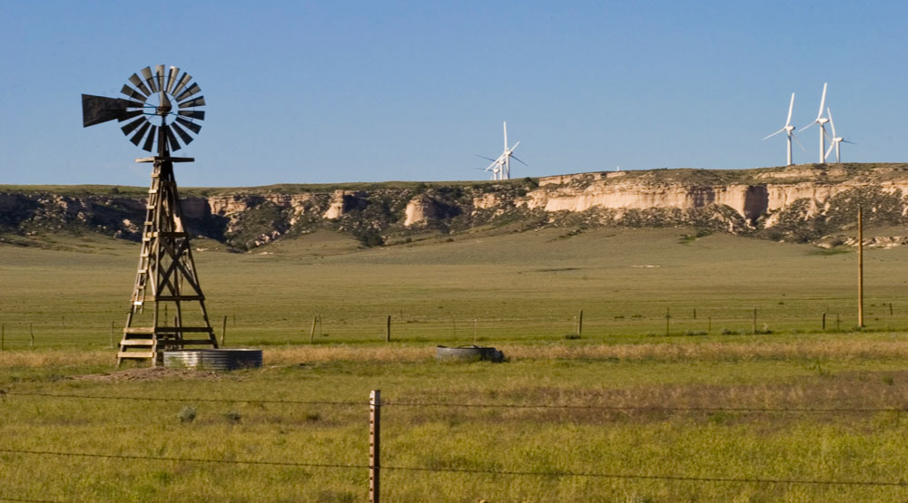 An old-school windmill turns in the foreground, and across some fences and prairie land, there are several white wind turbines