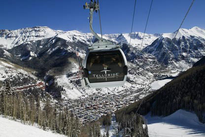 A gondola with Telluride's logo climbs its way up the cables away from the downtown area. Above town, blue mountain peaks are covered in snow.