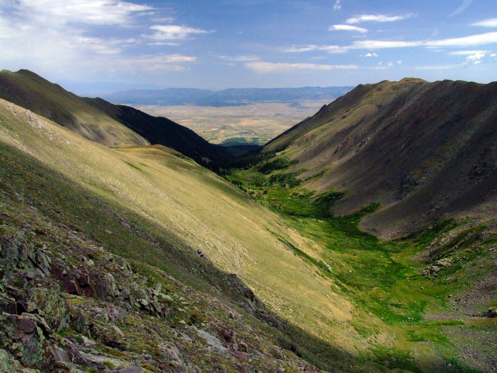 A verdant green valley lies between two barren, steep slopes near Westcliffe, Colorado. Clouds above cast shadows along parts of the valley and mountains.