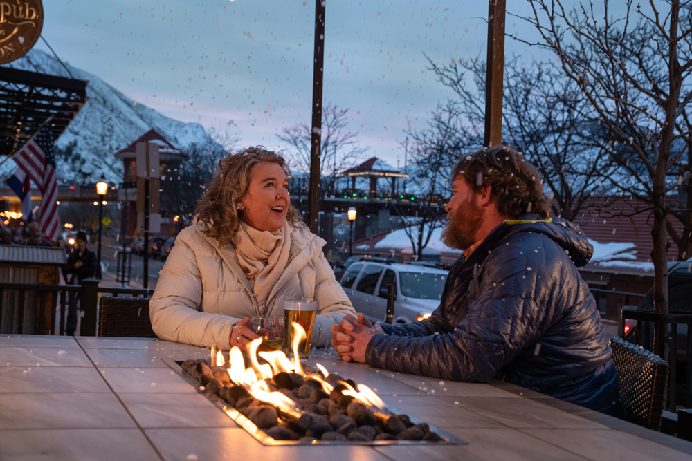 A couple sits at a table with a small firepit and snowy mountains outside