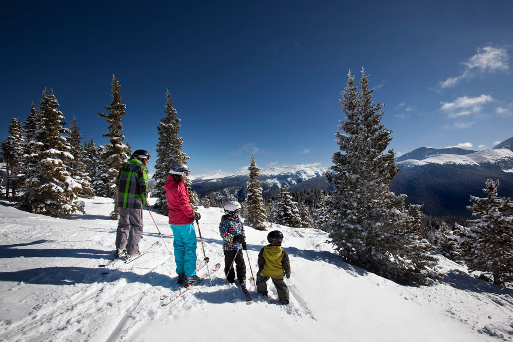 A family of two adult and two small children take a break from skiing and survey the scene of snow-covered mountains and tree on a ski trail in Winter Park, Colorado.